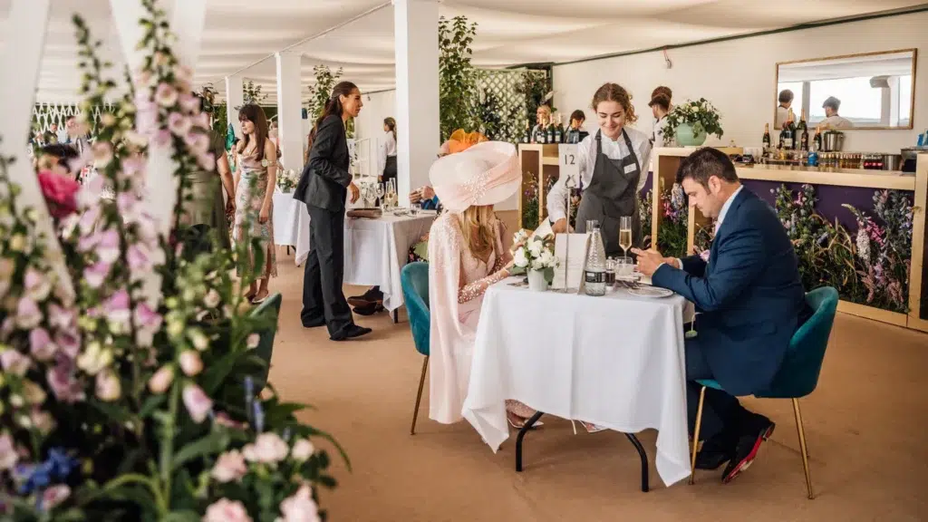 Elegant fine dining at The Deck, Royal Ascot, with guests dressed in stylish race-day attire enjoying gourmet cuisine in a luxurious floral-themed setting