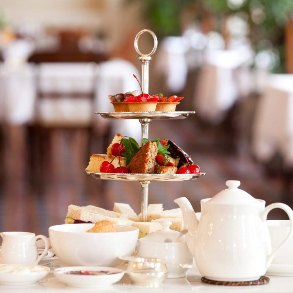 Royal Ascot Wyndham Restaurant – Elegant afternoon tea setup with a tiered cake stand featuring pastries, sandwiches, and desserts, accompanied by a classic white teapot and fine china in a refined dining setting