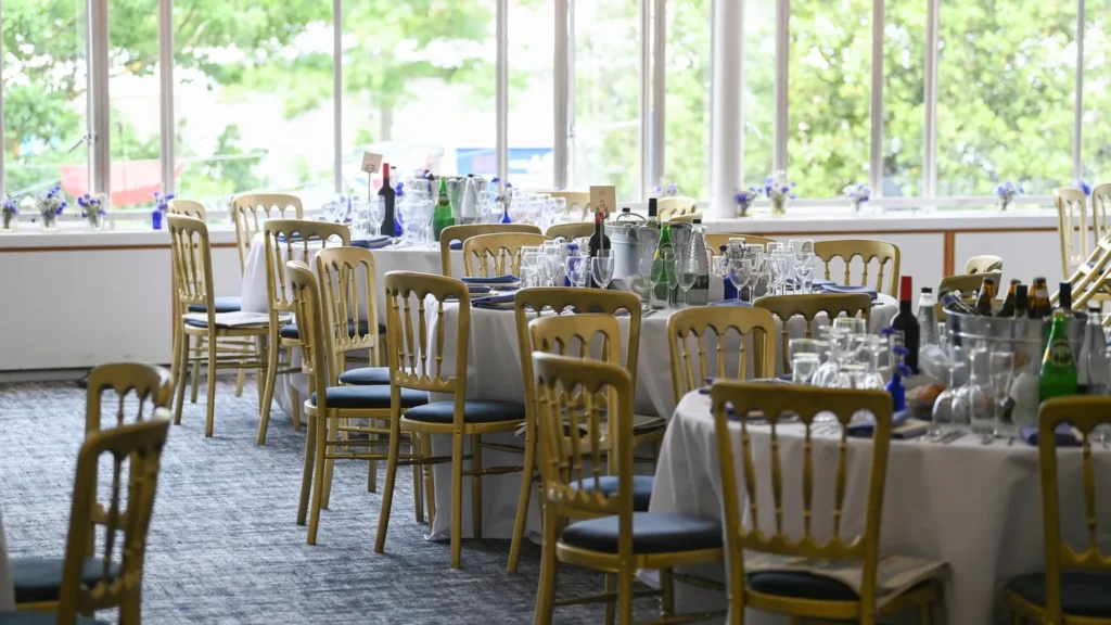Elegant dining setup at the Pavilion Restaurant, Royal Ascot, featuring gold chairs, round tables adorned with fine glassware, and a bright, airy ambiance with scenic outdoor views