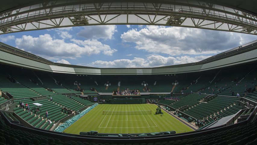 Wimbledon Centre Court under a partially open roof, showcasing its iconic grass court, tiered seating, and stunning sky backdrop