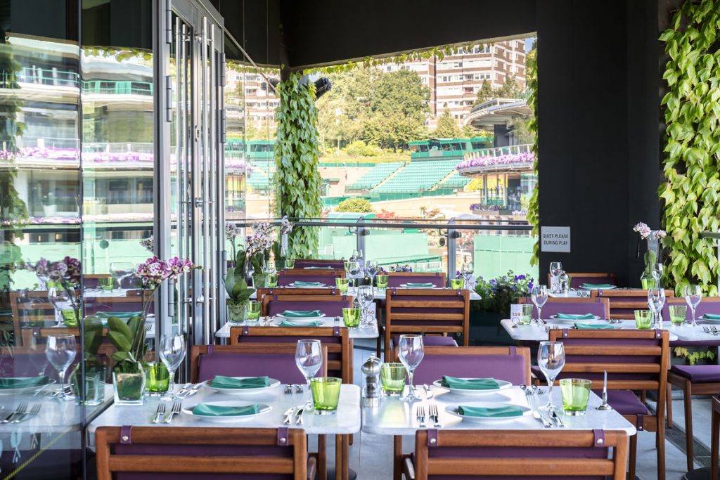Elegant courtside dining at Wimbledon with a view of lush green tennis courts, stylish table settings, and floral decor