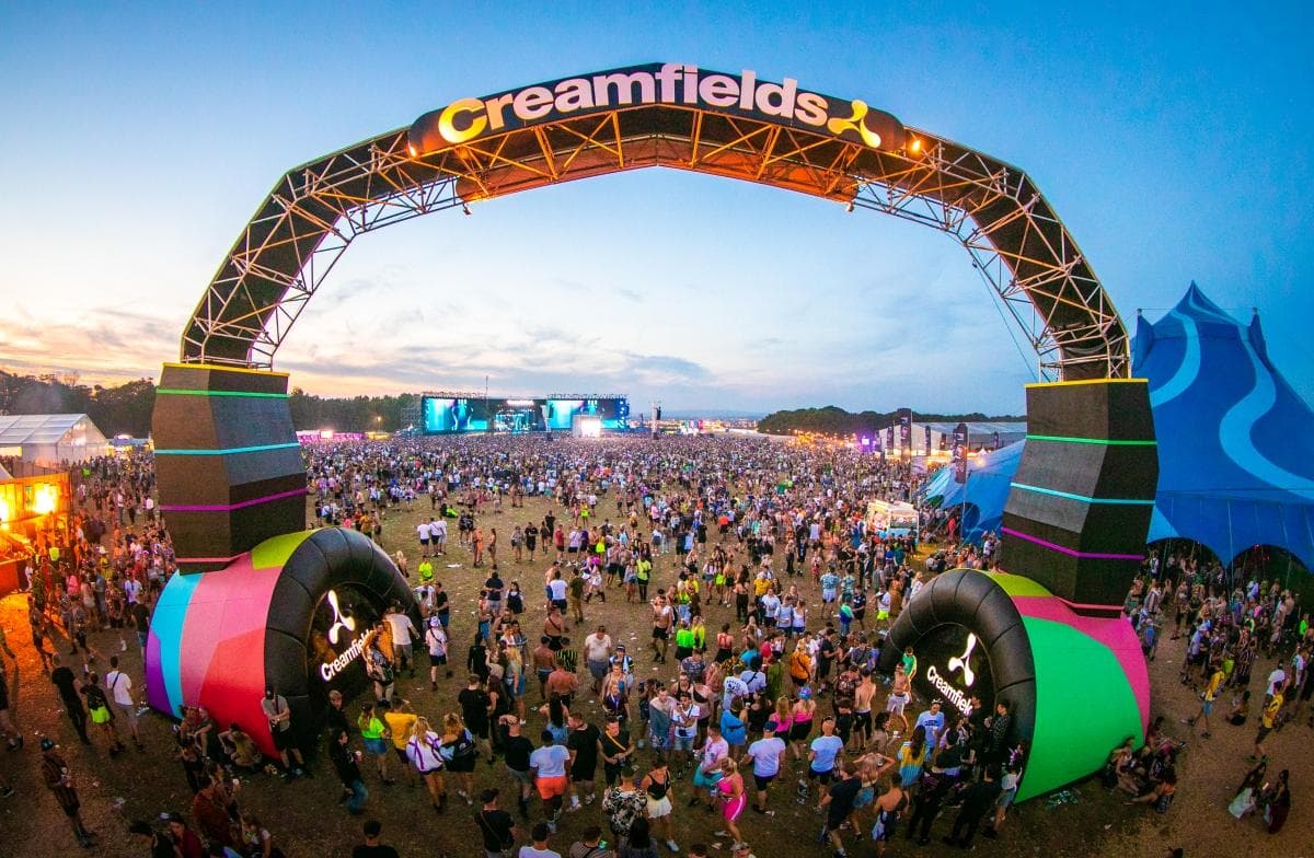 Massive crowd at Creamfields music festival under iconic entrance arch during sunset, with live stage and tents in the background