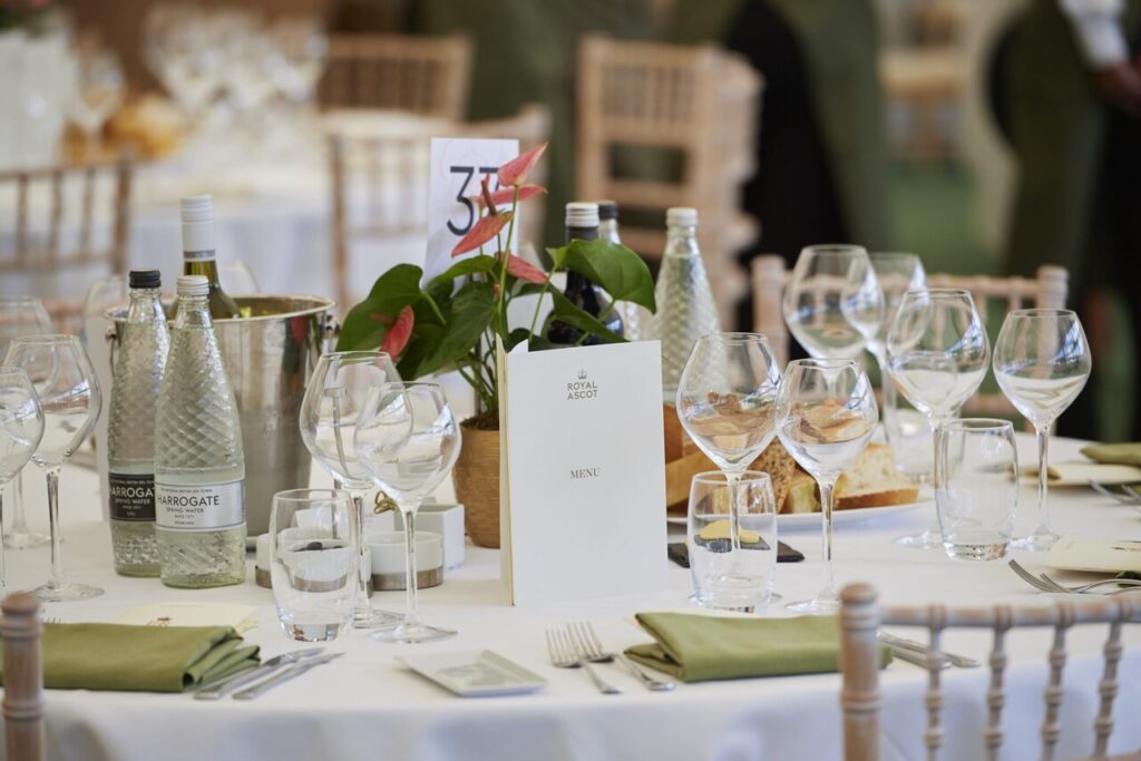 Elegant table setting at The Lawn Club, Royal Ascot, featuring fine glassware, premium bottled water, a chic floral centerpiece, and a luxury dining menu