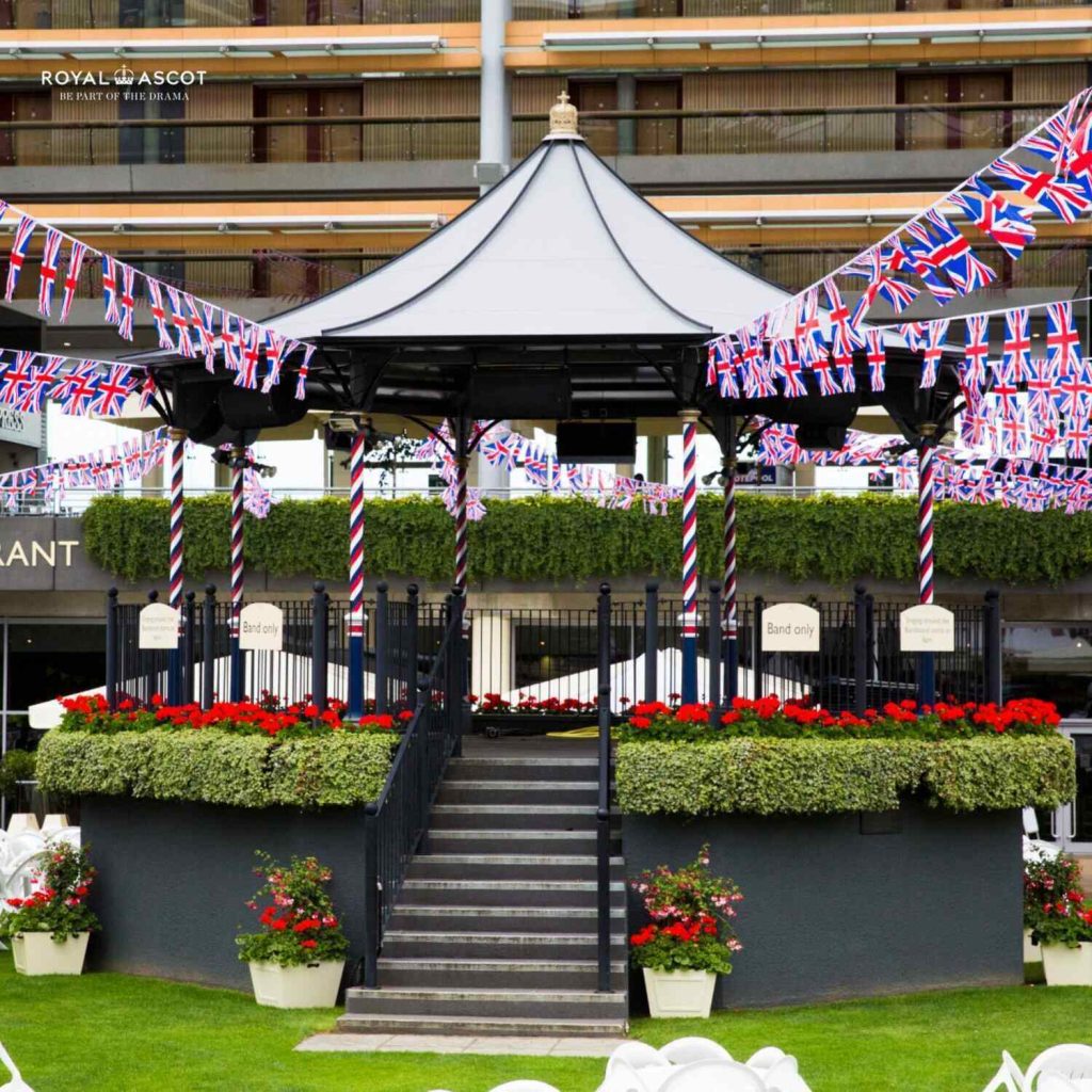 Royal Ascot Bandstand decorated with Union Jack bunting, surrounded by lush greenery and red floral arrangements, creating a festive atmosphere for spectators