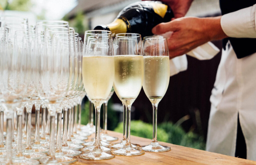 Royal Ascot hospitality – A waiter elegantly pouring champagne into sparkling flute glasses, set on a wooden table, creating a luxurious and celebratory atmosphere at the prestigious horse racing event