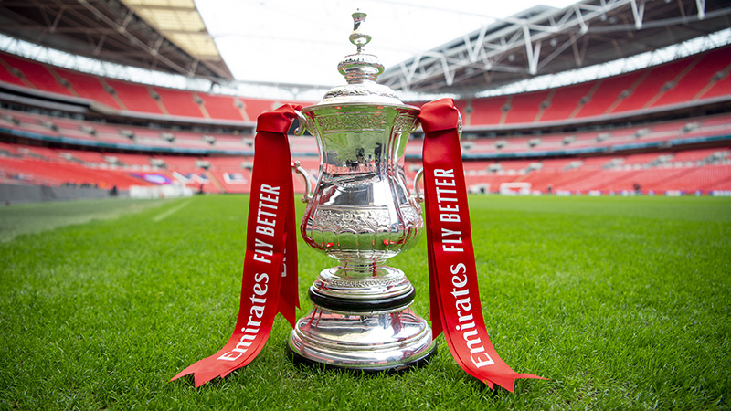 The FA Cup trophy displayed on the pitch at Wembley Stadium, featuring Emirates Fly Better red ribbons and the stadium’s iconic stands in the background.