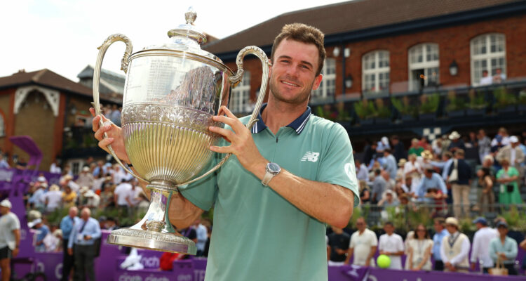 Tommy Paul, A tennis player smiles while holding the large silver LTA Championships trophy aloft, standing in front of a cheering crowd at an outdoor grass court venue