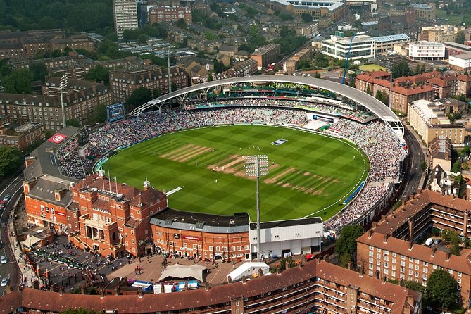 A panoramic view of The Kia Oval cricket ground, filled with spectators watching a match under a clear sky, with the historic pavilion and modern stands visible in the background