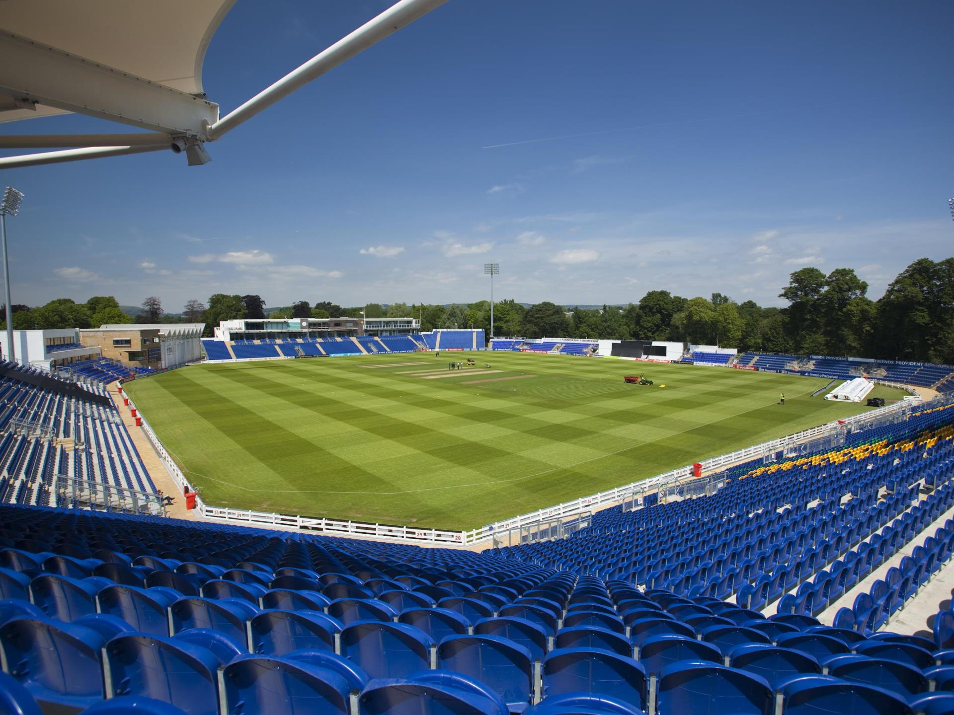 Wide-angle view of Sophia Gardens cricket stadium in Cardiff, featuring a well-maintained pitch, blue seating, and clear skies, with trees surrounding the ground