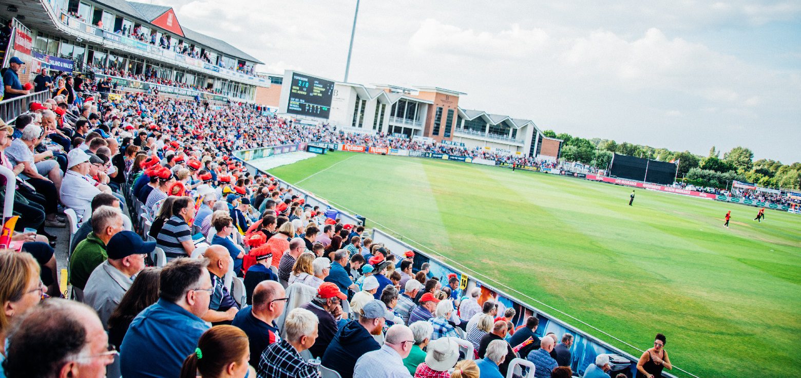 A lively crowd fills the stands at the Seat Unique Riverside cricket ground on a sunny match day, with players visible on the pitch and the modern pavilion in the background
