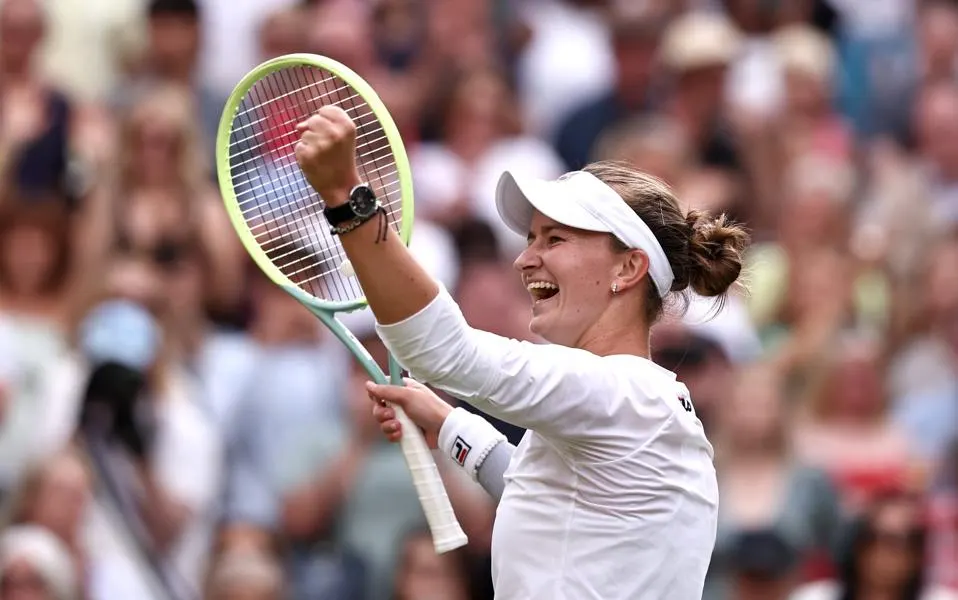 Female tennis player celebrates victory at Wimbledon, smiling and raising her fist with a crowd cheering in the background