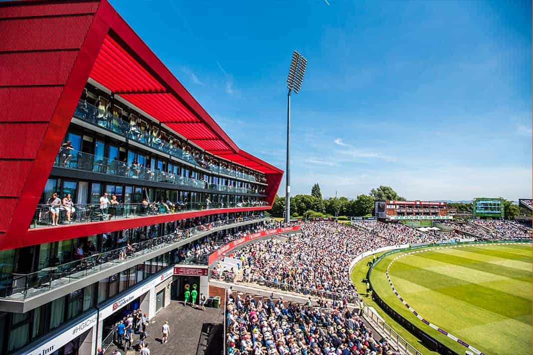 A packed Emirates Old Trafford cricket stadium during a sunny England vs India match, with fans filling the stands and a striking red hospitality building on the left