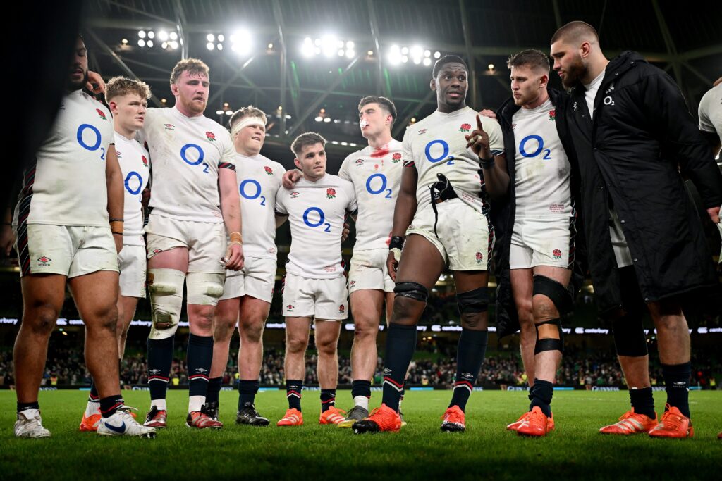 England rugby players in their white O2 sponsored kit huddle on the pitch during a Six Nations match, with stadium lights and crowd in the background.