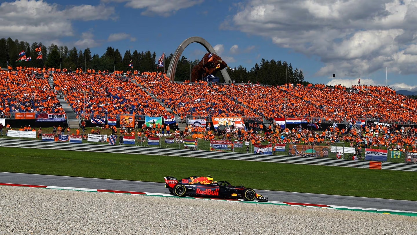 A Red Bull Formula 1 car races past a packed grandstand of fans in orange at the Austrian Grand Prix, with the iconic bull statue and forested hills in the background