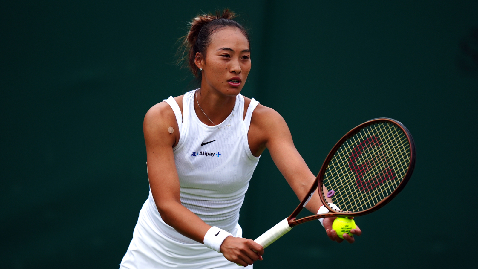 Zheng Qinwen prepares to serve during a Wimbledon match, wearing a white sleeveless top and holding a tennis ball and racket on a grass court
