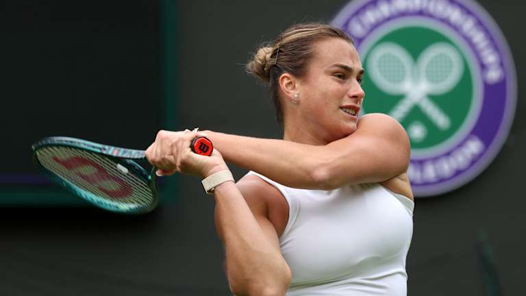 A female tennis player, wearing a white outfit, follows through on a backhand shot at Wimbledon, with the tournament’s iconic green and purple logo visible in the background.