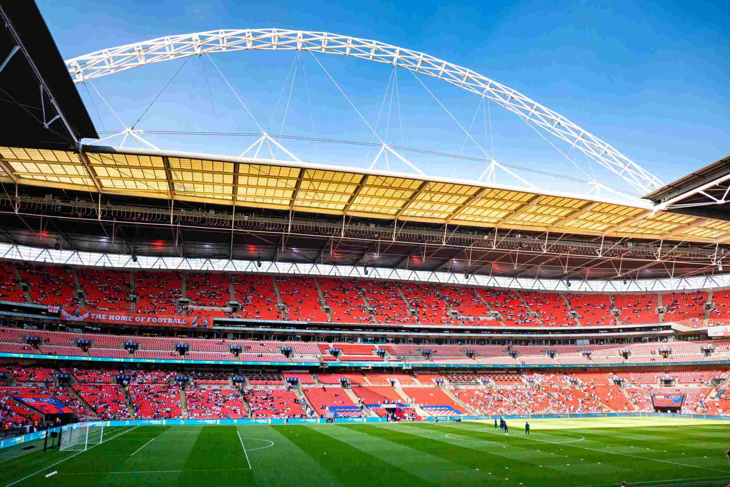 This image shows Wembley Stadium in London, one of the most iconic football venues in the world. It features the distinctive arch that spans over the stadium, as well as the red seating and expansive three-tier stands. A banner that reads "THE HOME OF FOOTBALL" can be seen, affirming Wembley’s central place in English football culture
