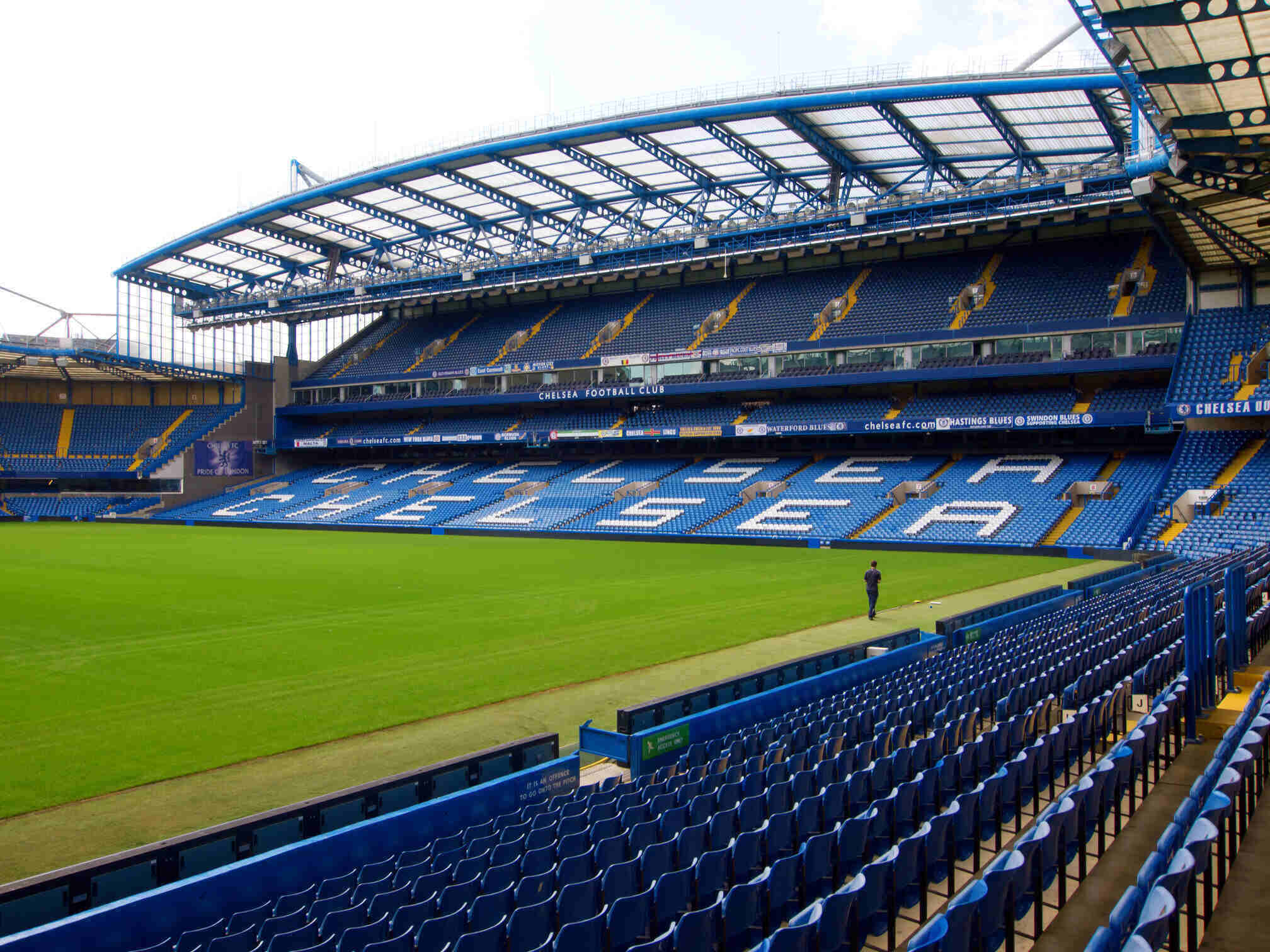 This image shows Stamford Bridge, the iconic home stadium of Chelsea Football Club in London. The empty stands prominently display the word "CHELSEA" in white seating, set against the blue seats that reflect the club's primary color. The stadium features a modern design with a partial roof and multiple tiers