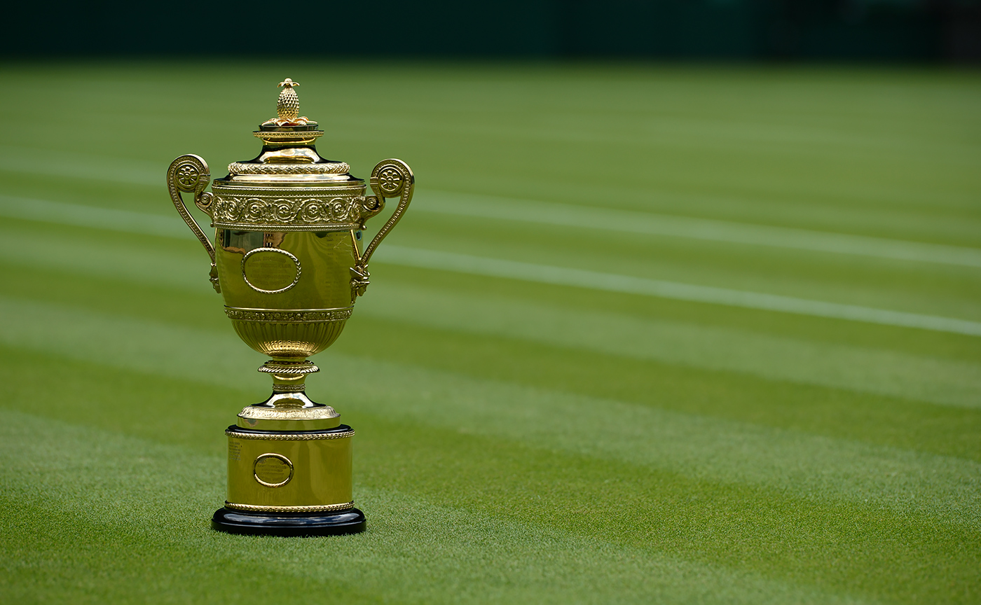 The Wimbledon Gentlemen’s Singles Trophy sits on the pristine grass court, gleaming gold under natural light, with clean white lines marking the field in the background