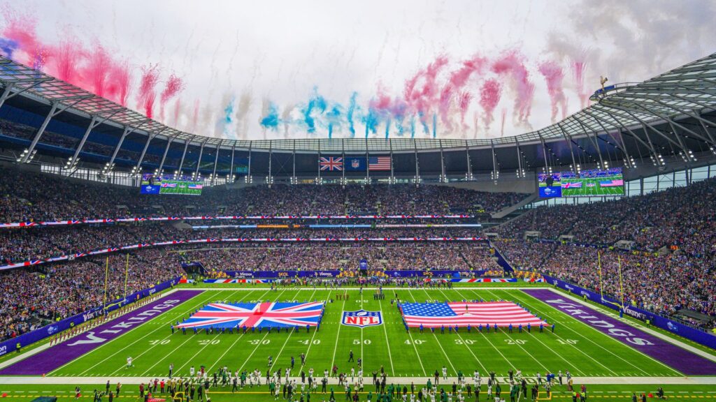 NFL London game at Tottenham Hotspur Stadium, featuring the UK and US flags on the field with fireworks overhead and a packed crowd in attendance.