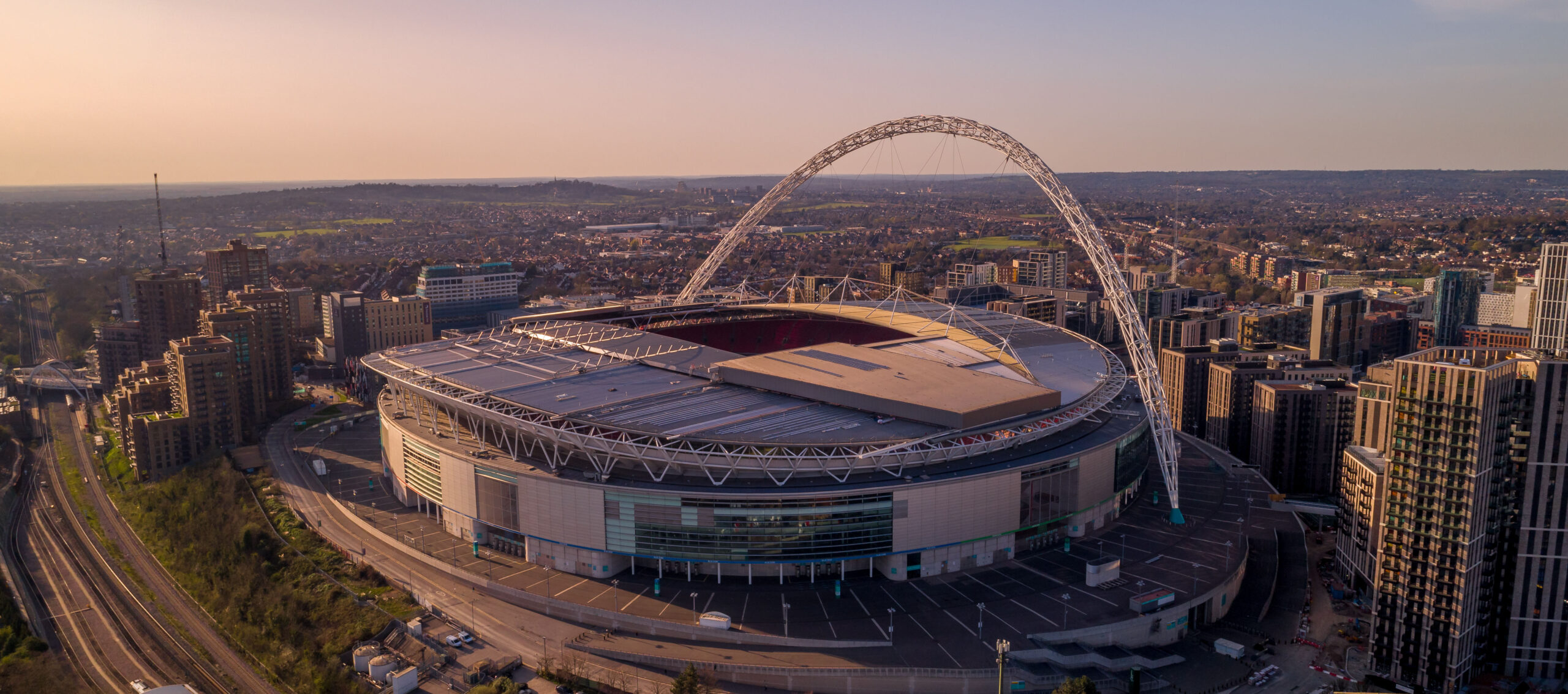Aerial view of the iconic Wembley Stadium and its famous arch at sunset, overlooking the London skyline, a premier venue for world-class sports and music hospitality.